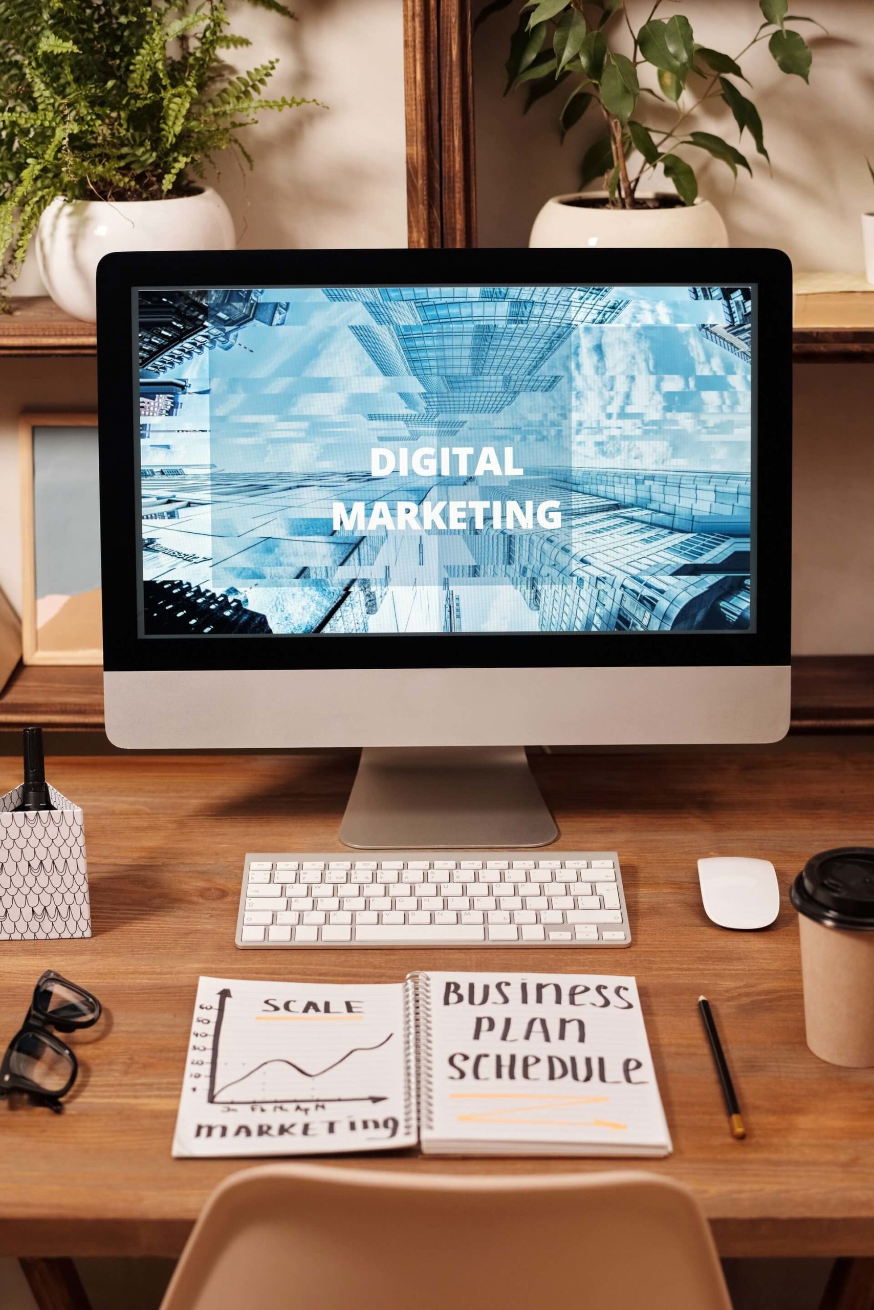 digital-marketing Desktop computer on a wooden table with a notebook, cup, pen, glasses and plants in a white round vase.