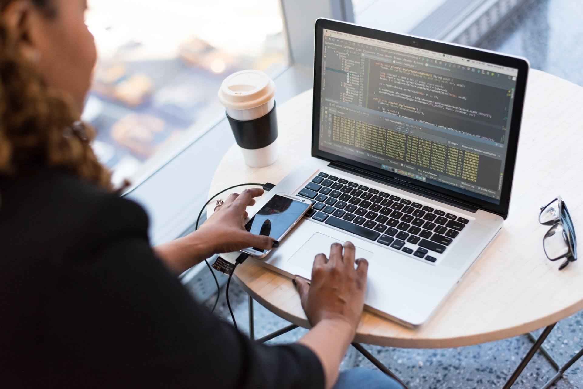 web-development A person using a laptop with a mobile phone, eyeglasses, and a paper cup on top of a round table.