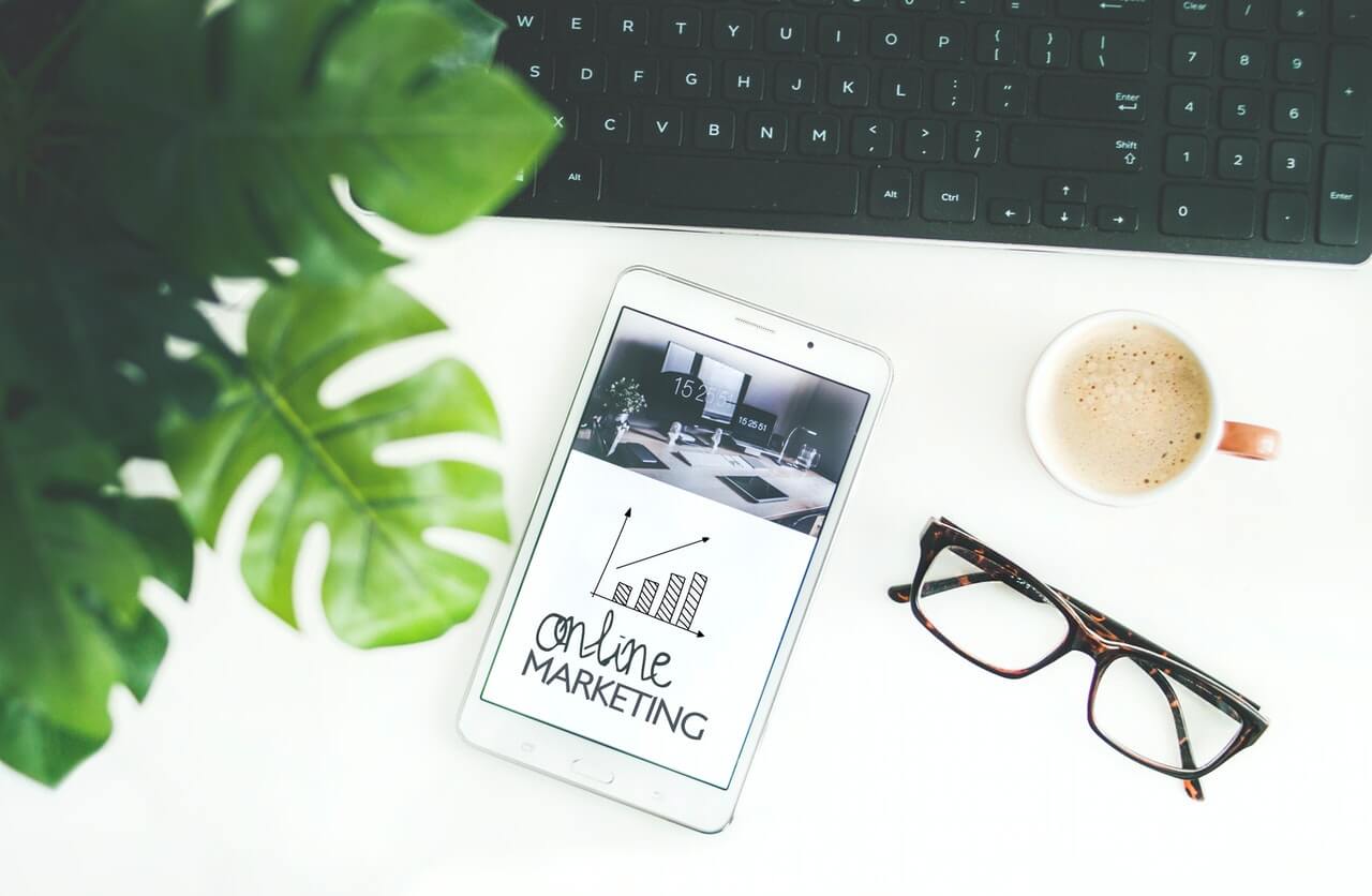 services-digital-marketers White table with a green leaf, black keyboard, tablet, glasses, and a mug of coffee on top.