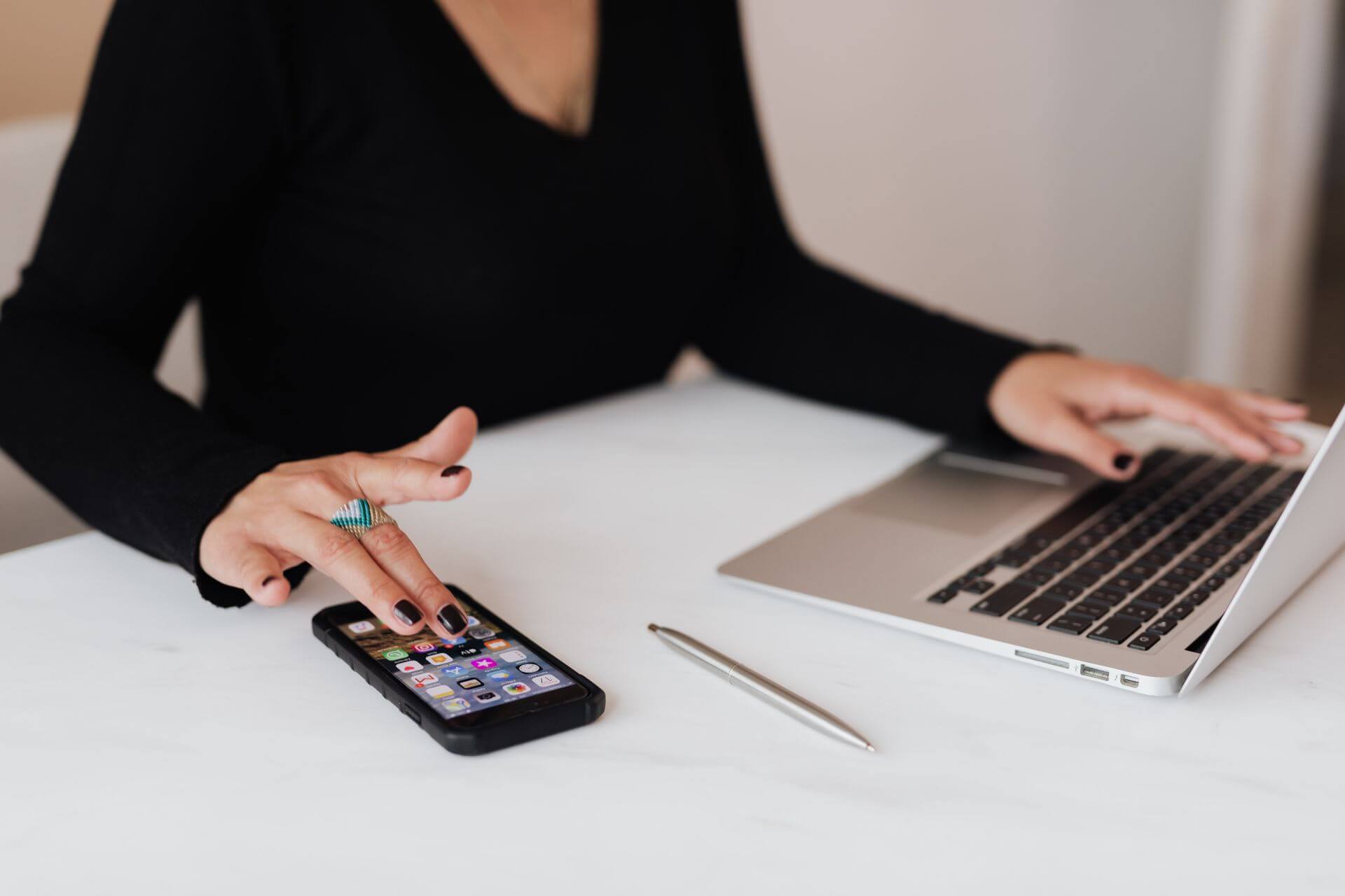 Benefits of Using Social Media for Small Businesses A person wearing a black long-sleeved shirt, using a mobile phone and laptop with a pen in the table at Smart Works Intl.