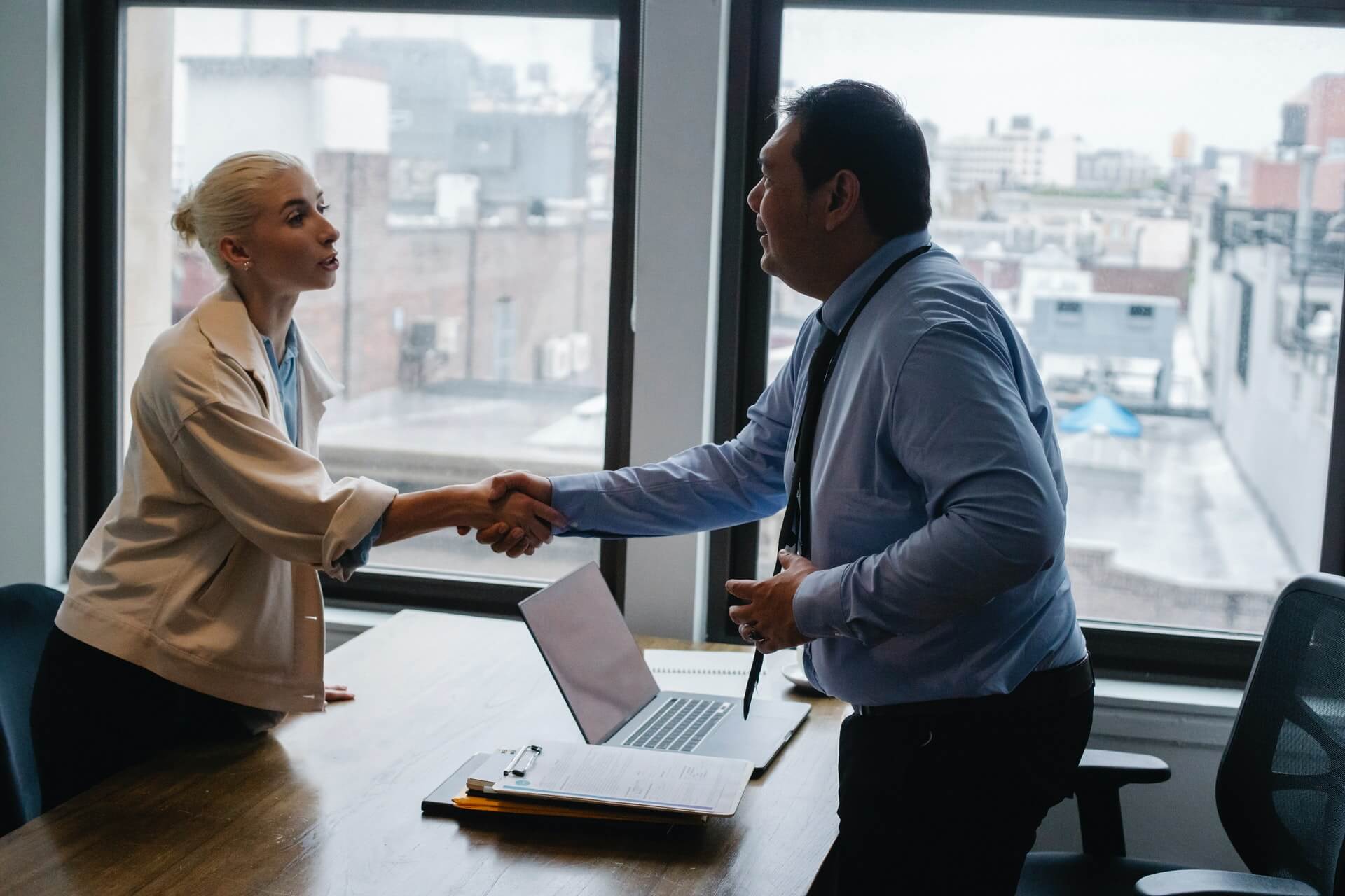 Get Great Hires For Your Small Business min A man and a woman wearing formal attire shaking hands in the office with a laptop and a clip board on the table