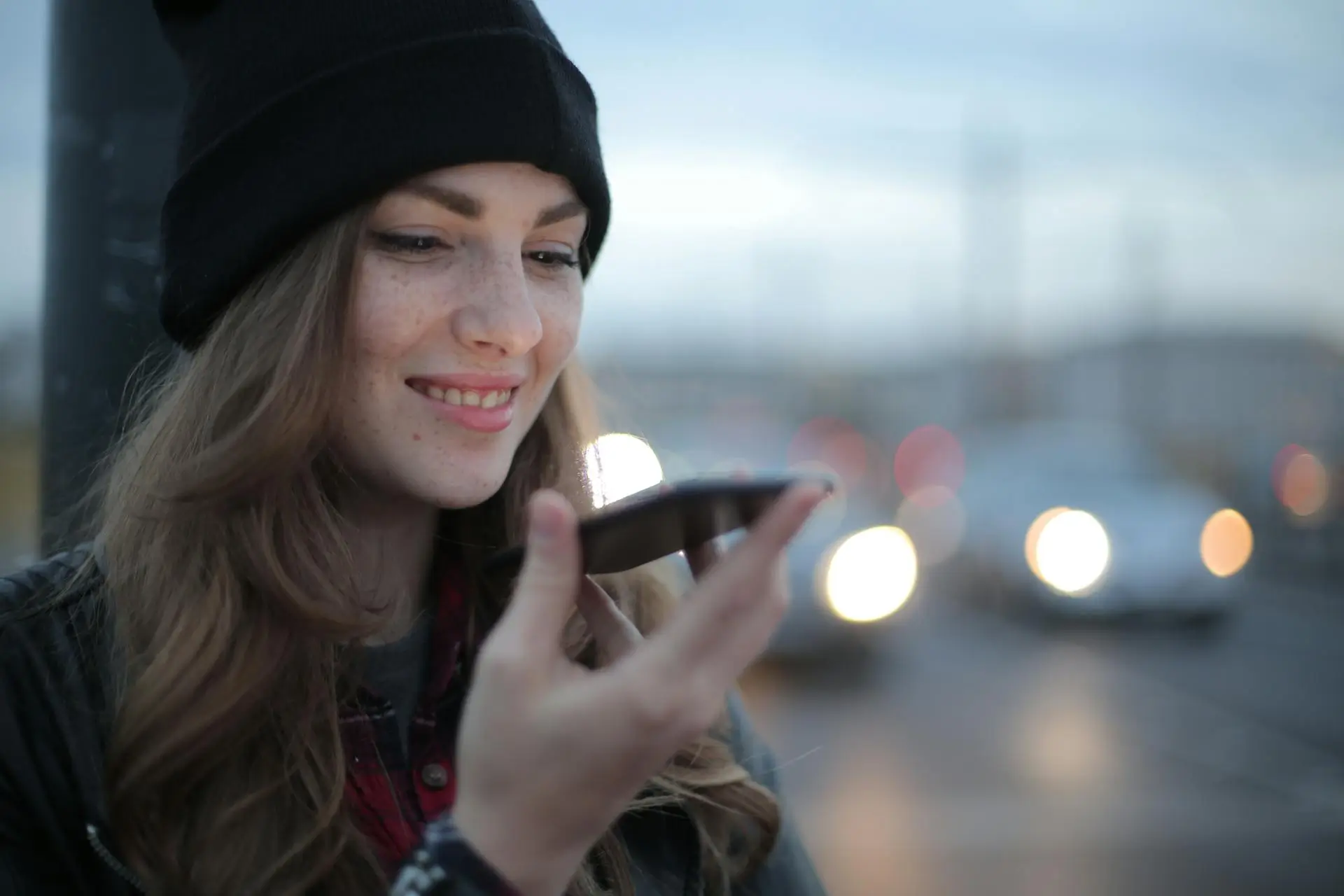 Staying Ahead with Voice Search Optimization A woman smiling, with a blonde hair, wearing bonnet, black leather jacket, while holding a phone. Using voice search optimization